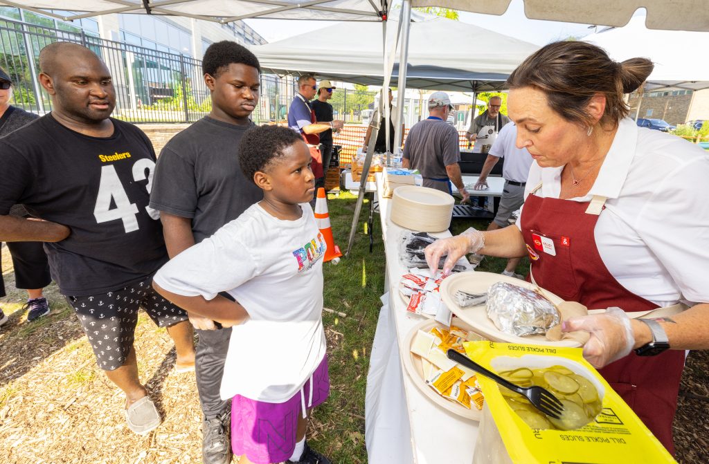 Volunteer from Tyson Foods serves burgers and toppings to a young guest in line at the Ronald McDonald House Summer BBQ Reunion.