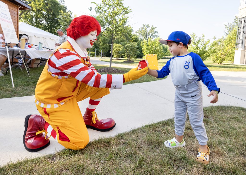 Ronald McDonald hands a Happy Meal to a young boy in a baseball uniform at the Summer BBQ Reunion.